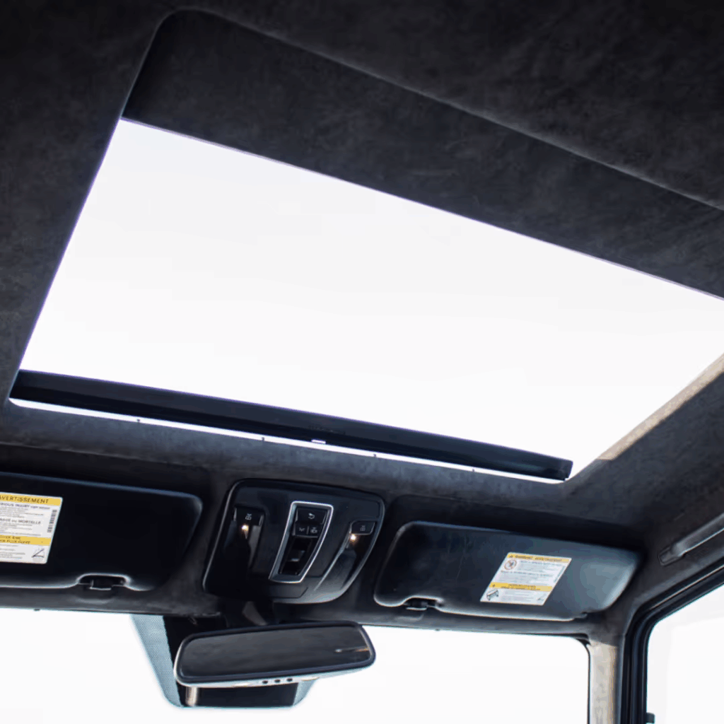 Interior view of a vehicle's panoramic sunroof, showcasing controls and overhead storage compartments, with a bright sky visible through the open sunroof.