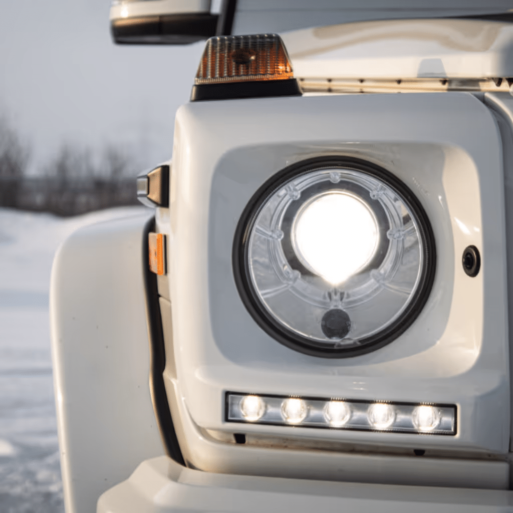 Close-up of a white vehicle's front headlight, featuring a round main light and LED strips below, set against a snowy landscape.