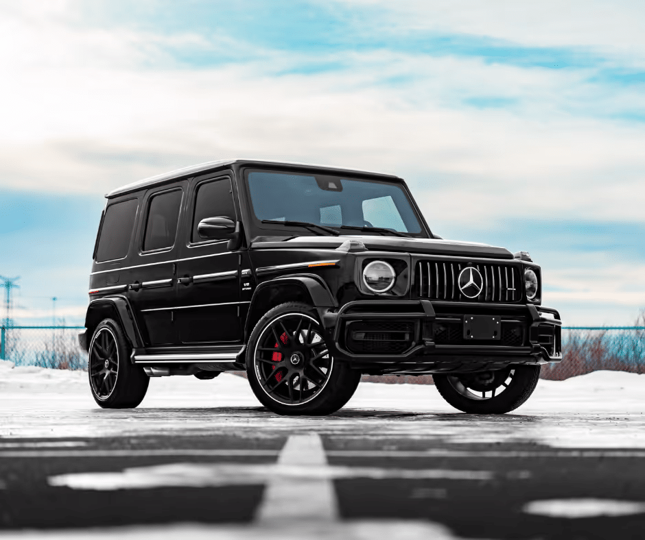 A black Mercedes-Benz G-Class SUV parked on a snowy surface, showcasing its luxurious design and bold styling, with visible alloy wheels featuring red brake calipers and a cloudy blue sky in the background.