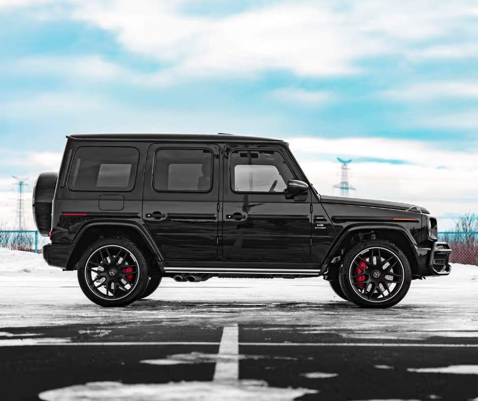 Black Mercedes-Benz G-Class SUV parked on a snowy surface, showcasing its sleek design, prominent wheel arch, and red brake calipers against a backdrop of a cloudy sky.
