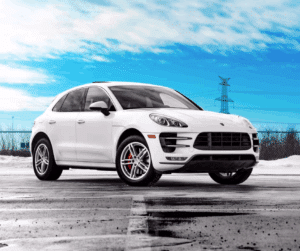 White Porsche Macan parked on a wet asphalt surface with a bright blue sky in the background, showcasing its sporty design and performance-oriented features.
