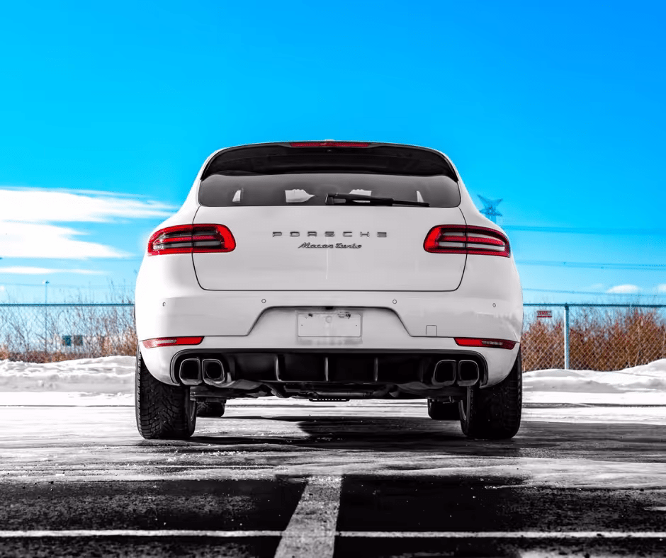 White Porsche Macan parked in a snow-covered lot, showcasing its sleek rear design and dual exhaust pipes against a bright blue sky.