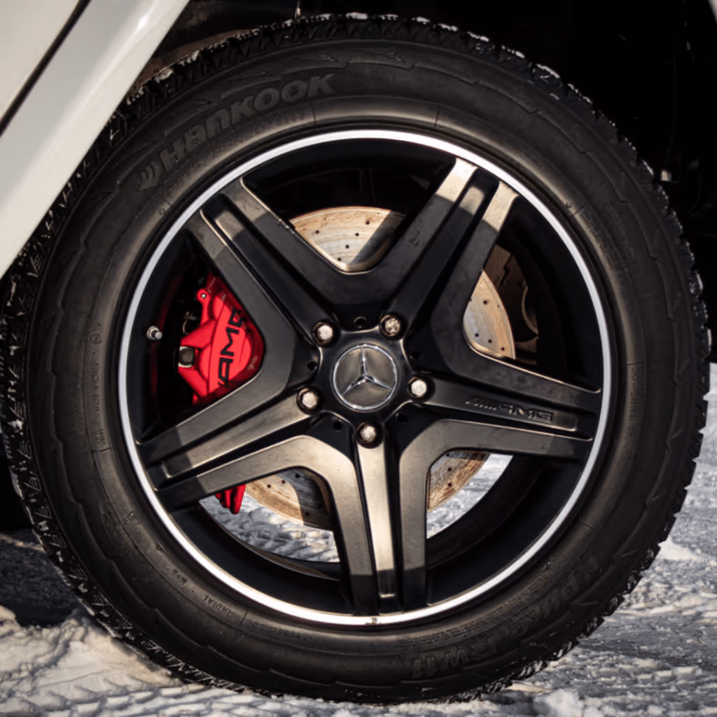 Close-up of a black Mercedes AMG wheel featuring distinctive five-spoke design, red brake caliper, and Hankook tire, set against a snowy background.