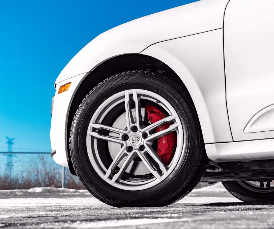 Close-up of a white car's wheel showcasing sleek alloy rims, red brake calipers, and a snowy background under a bright blue sky, emphasizing performance and design.