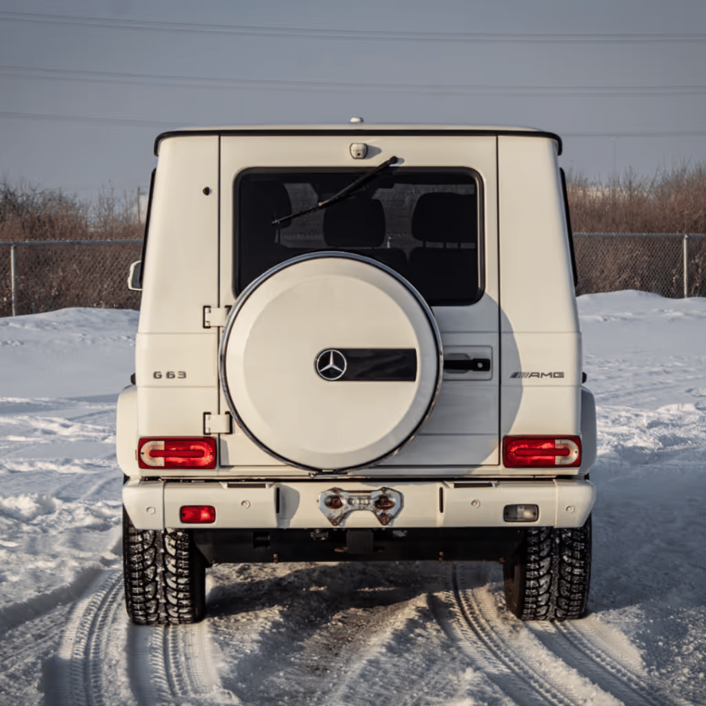 A white Mercedes-Benz G63 AMG parked on snow, showcasing its rear design, spare tire cover, and distinctive taillights. The vehicle is surrounded by a snowy landscape, emphasizing its rugged, off-road capabilities.