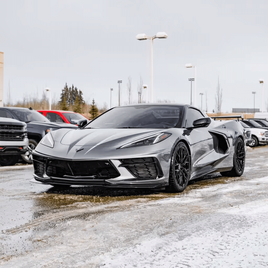 Sleek silver Chevrolet Corvette C8 parked in a dealership lot, showcasing its aerodynamic design and glossy finish, surrounded by other vehicles on a snowy day.
