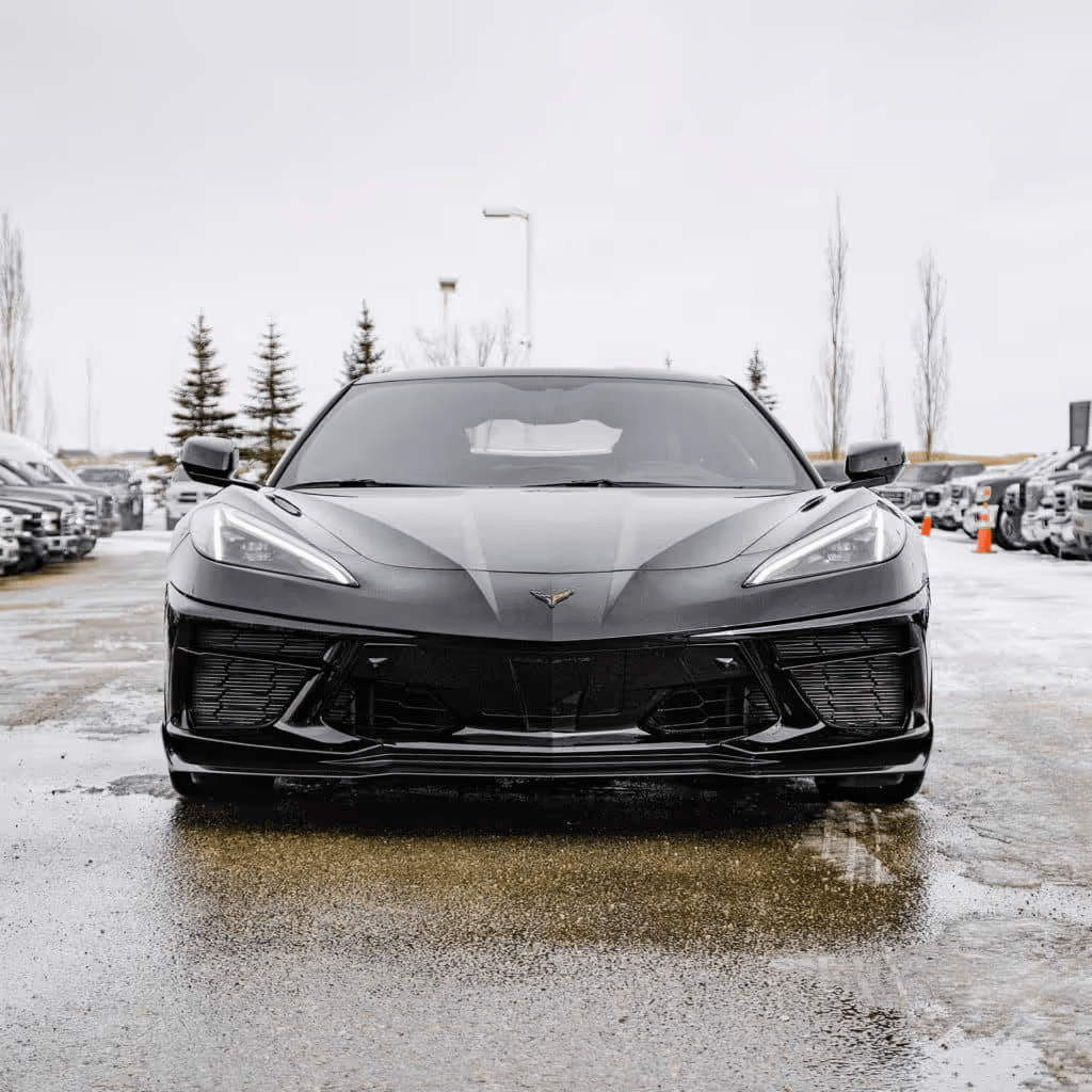 Front view of a black Chevrolet Corvette parked in a lot, showcasing its sleek design and aggressive stance on a wet surface with trees in the background. Ideal for automotive enthusiasts and car buyers.