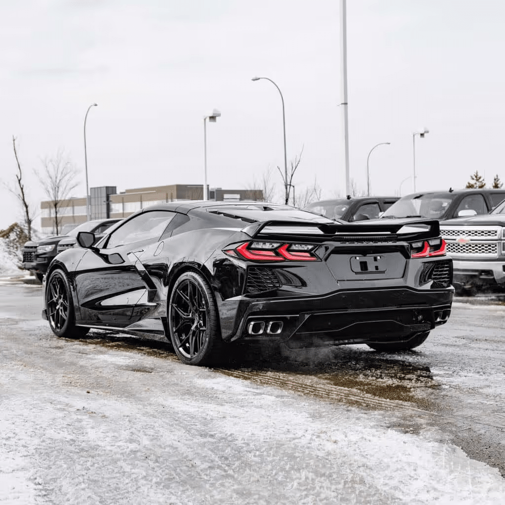 Black Chevrolet Corvette C8 parked in a snowy lot, showcasing a sleek rear design and distinctive taillights, surrounded by other vehicles.