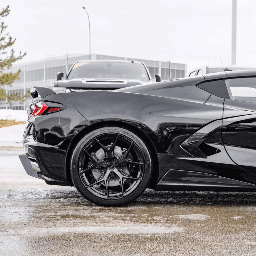 Black Chevrolet Corvette C8 parked outdoors, showcasing its sleek design and modern alloy wheels, with a blurred background of snow and a building.