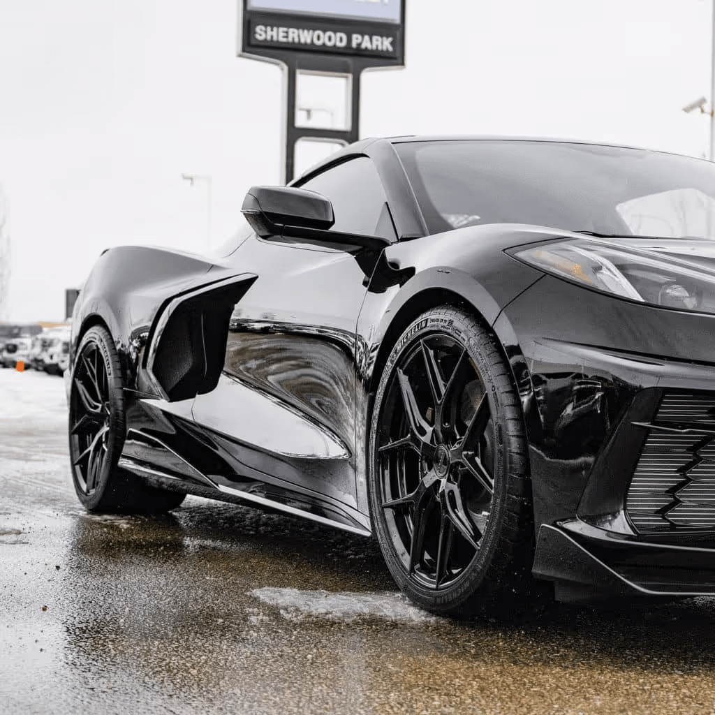 Black 2022 Chevrolet Corvette C8 parked outside a dealership in Sherwood Park, showcasing its sleek design, aerodynamic features, and sporty black alloy wheels, with a reflective surface on wet pavement under overcast skies.