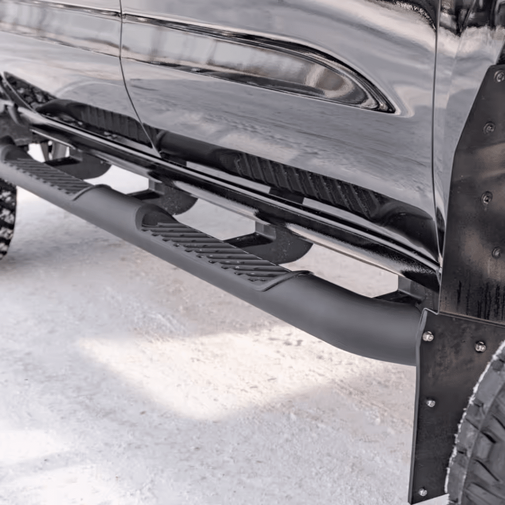Close-up of a black truck's side steps showcasing a rugged design, set against a snowy background. The durable material features a textured surface for enhanced grip and stability while entering or exiting the vehicle.