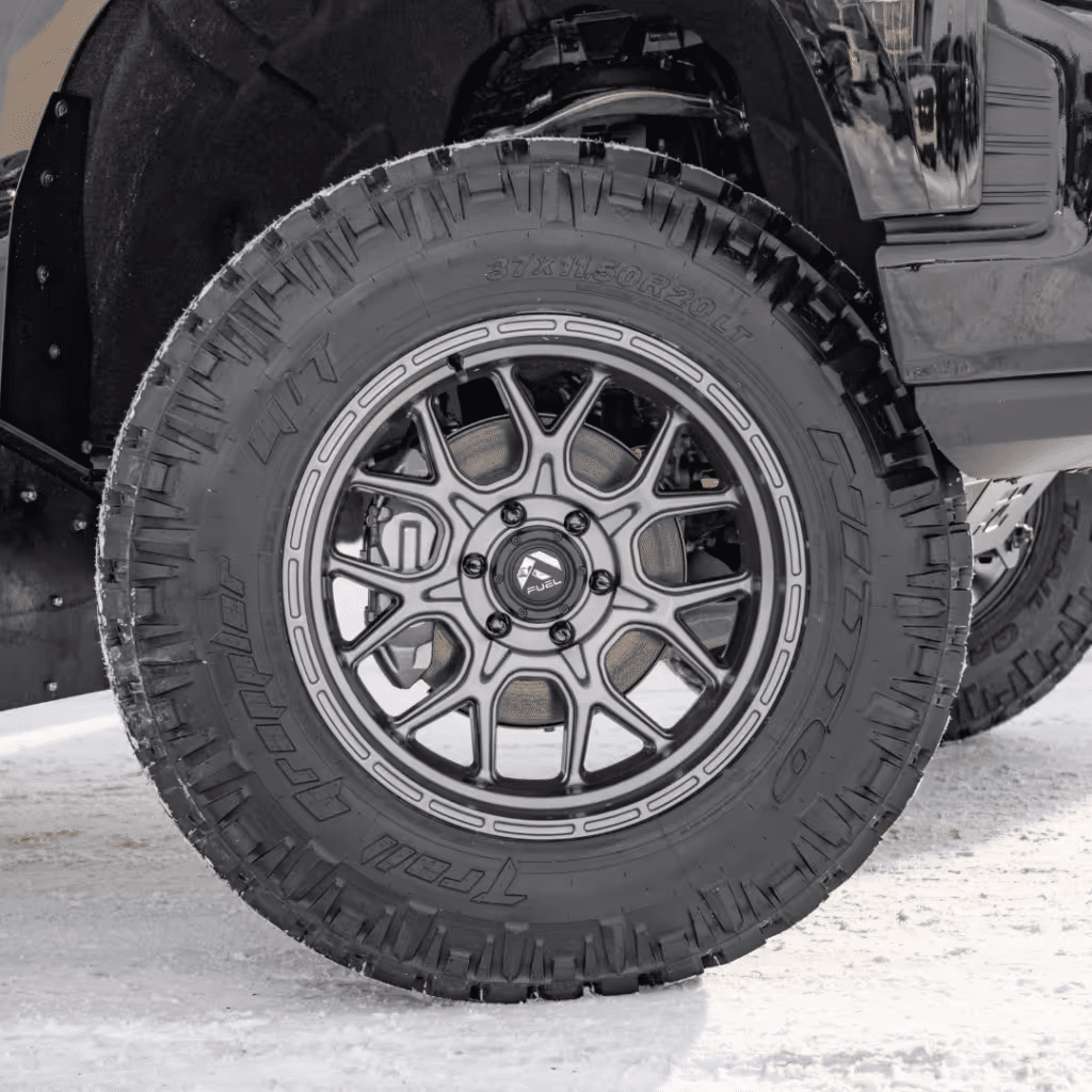 Close-up of a rugged all-terrain tire mounted on a black truck, showcasing a modern alloy wheel design and aggressive tread pattern, suitable for off-road conditions. The scene is set in a snowy environment, emphasizing the tire's durability and traction capabilities.