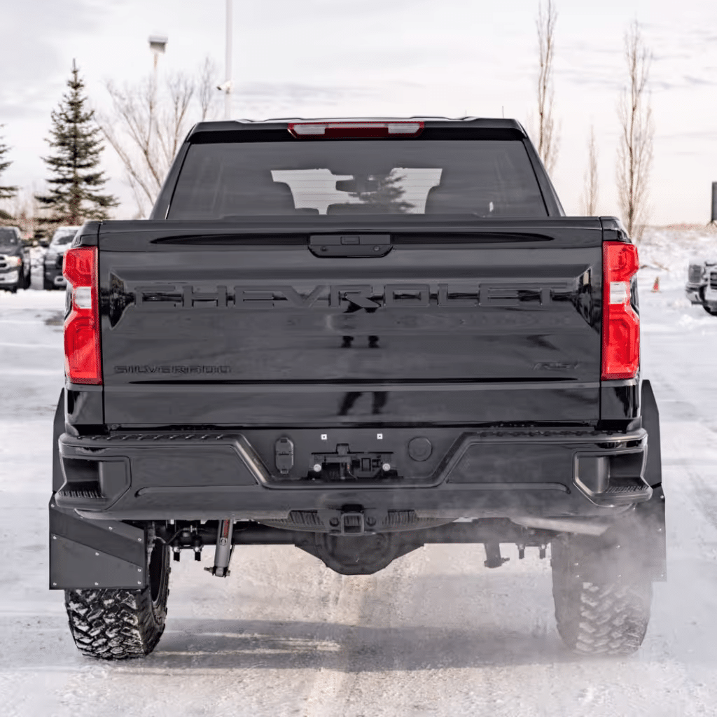 Rear view of a black Chevrolet Silverado truck parked in a snowy landscape, showcasing its bold design, prominent logo, and rugged tires. The scene captures the truck's sturdy build and off-road capabilities against a backdrop of winter trees and other vehicles.