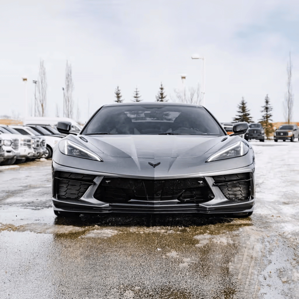 Front view of a sleek gray Chevrolet Corvette parked on a winter day, showcasing its aerodynamic design and modern styling with snow-dusted ground and trees in the background.