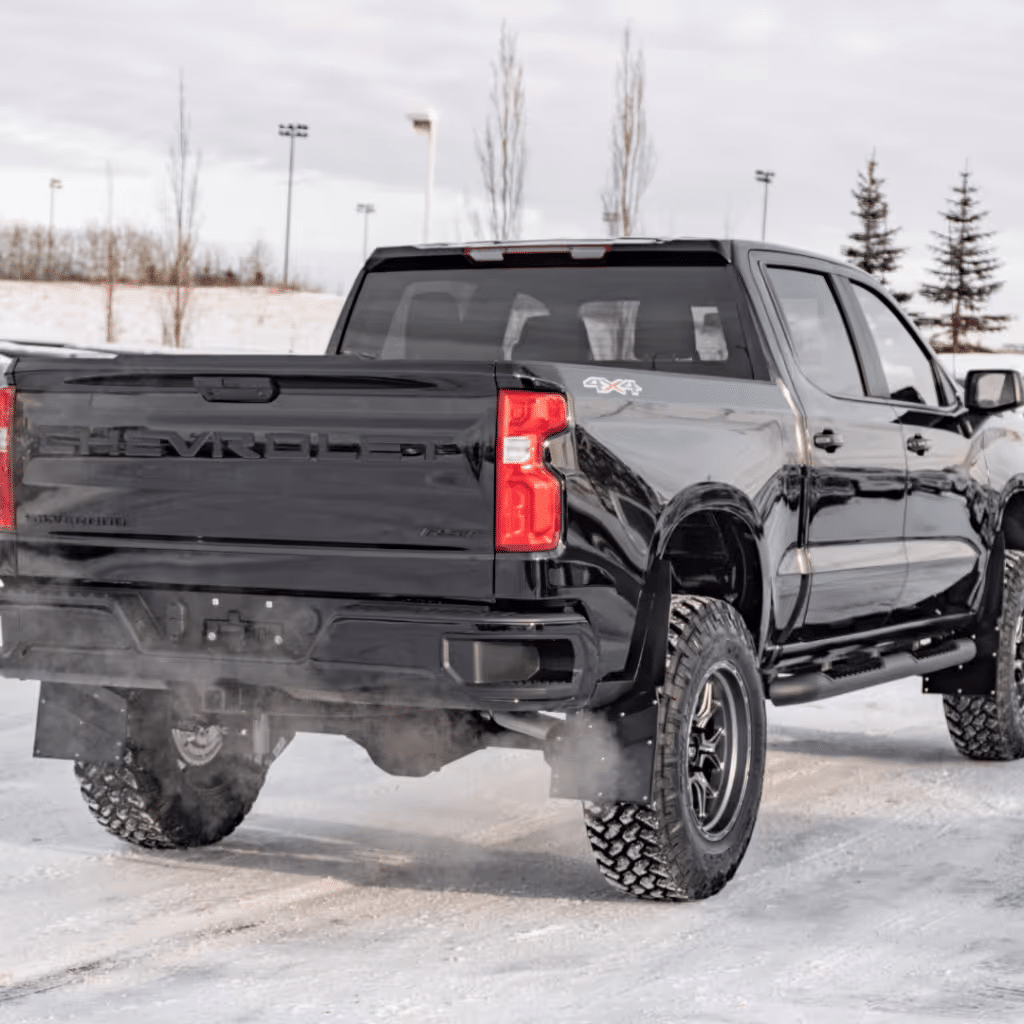 Black Chevrolet Silverado pickup truck parked on snowy terrain, showcasing its rugged design and off-road capabilities with visible tire tracks and a backdrop of trees and sports lights.