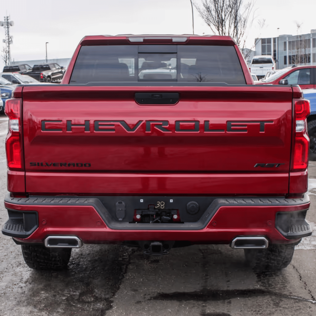 Red Chevrolet Silverado RST parked, showcasing its rear design, including large 'CHEVROLET' and 'SILVERADO' badges, tow hitch, and dual exhaust tips, with additional vehicles visible in the background.