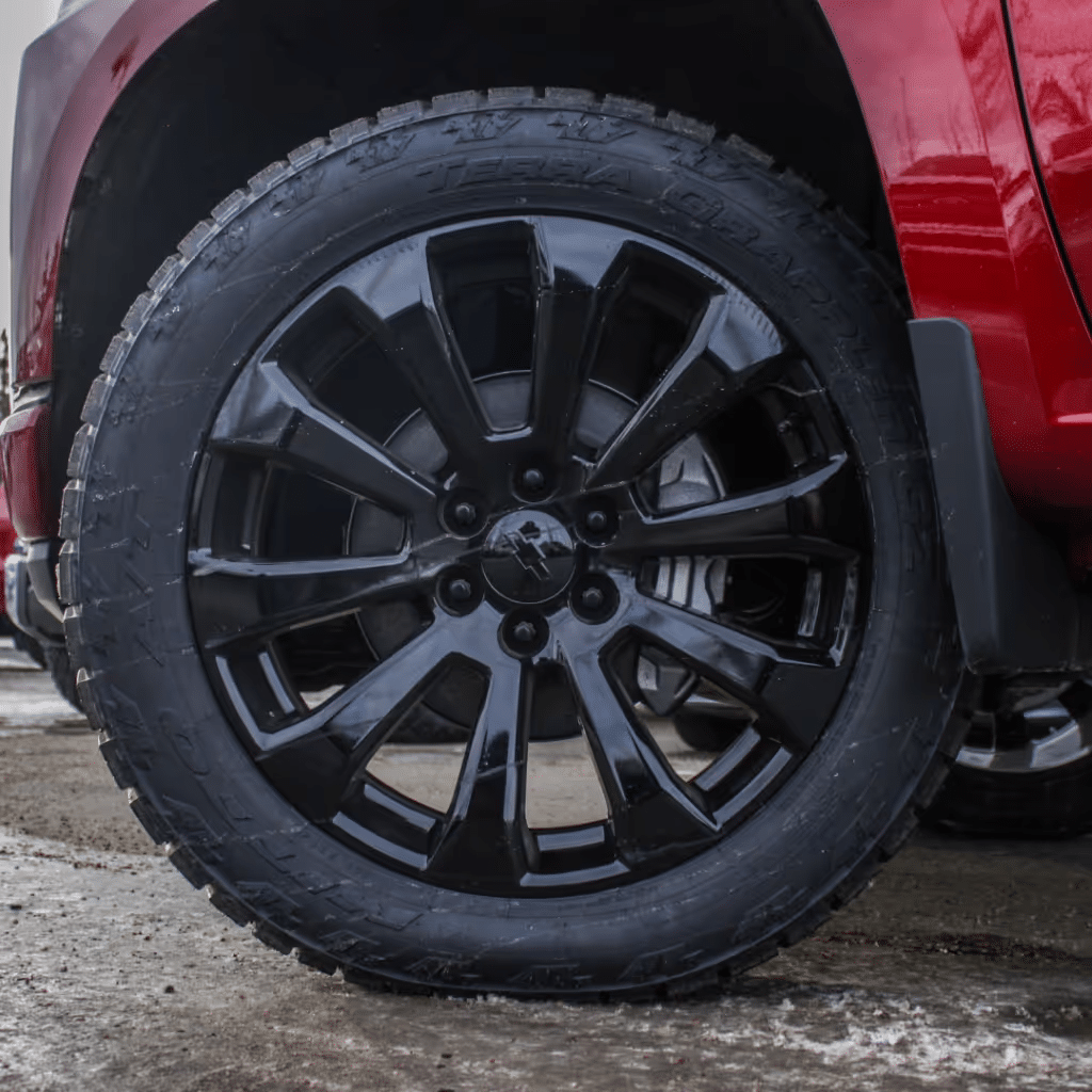 Close-up of a black alloy wheel with a rugged tire, showcasing a modern design, mounted on a red SUV parked on a wet surface. The wheel features a geometrically intricate pattern that enhances the vehicle's sporty appearance.