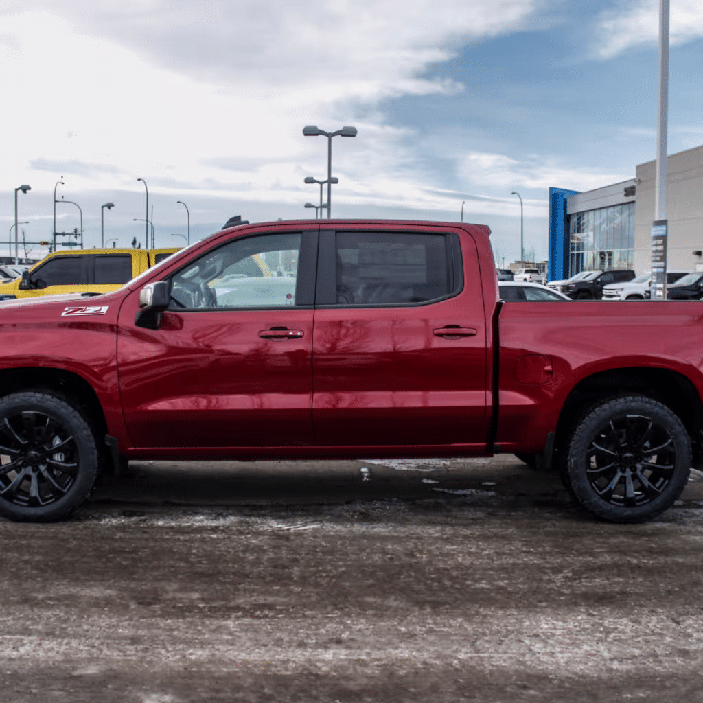 Side view of a red pickup truck with black rims parked at a dealership, showcasing its robust design and off-road capabilities against a cloudy sky.