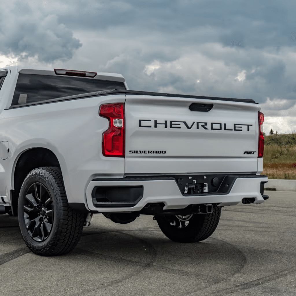 Rear view of a white Chevrolet Silverado RST pickup truck showcasing its sleek design, bold Chevrolet branding, and black alloy wheels, set against a cloudy sky.