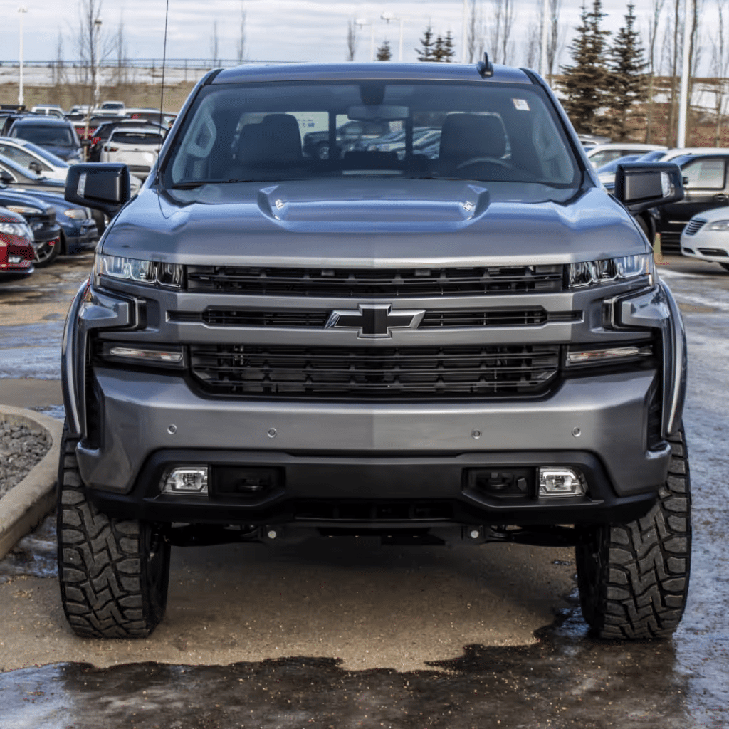 Front view of a grey Chevrolet Silverado truck showcasing its bold design, featuring a prominent grille, headlights, and rugged tires, parked in a dealership lot surrounded by other vehicles.