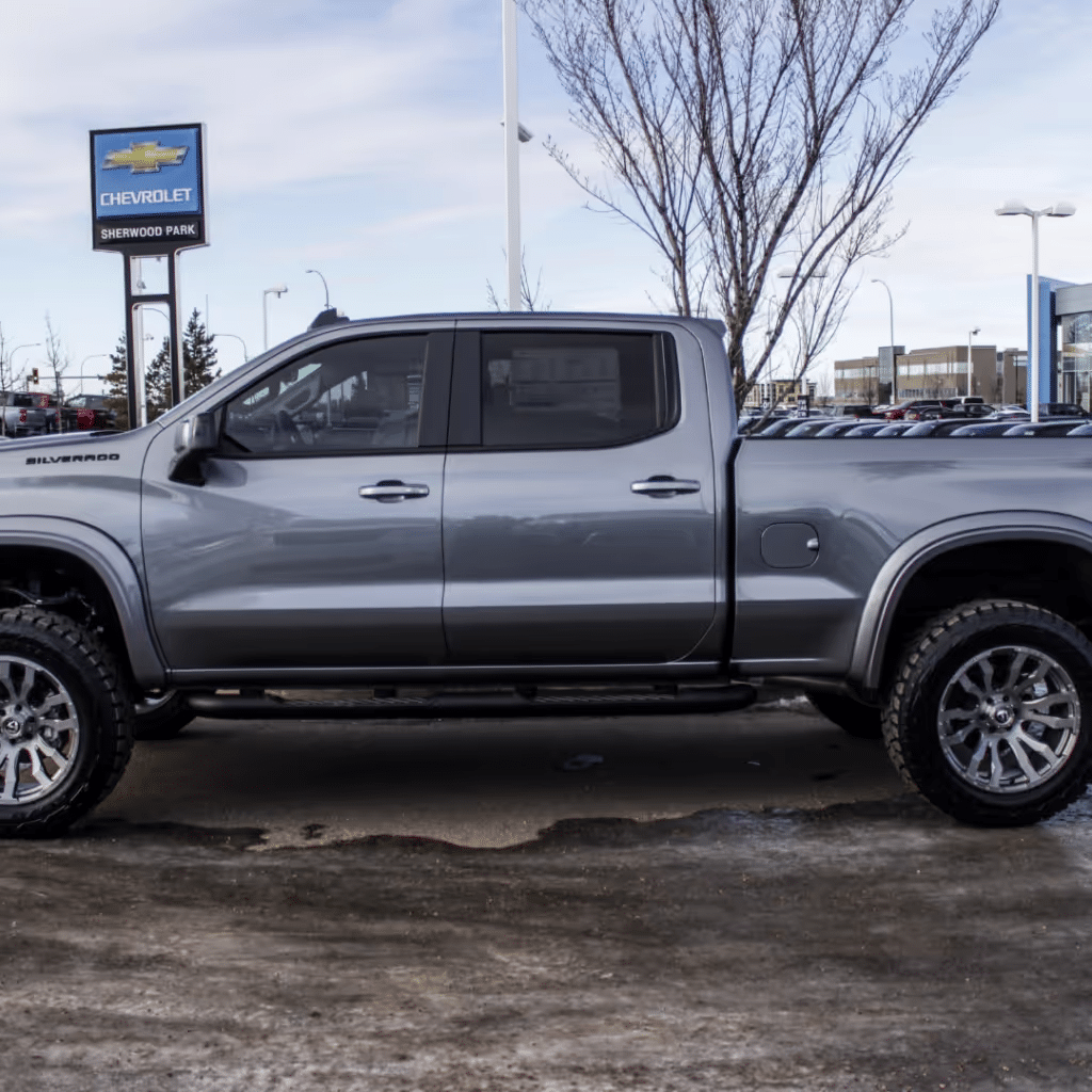 Silver Chevrolet Silverado pickup truck parked at a dealership, showcasing a modern design and off-road tires against a clear sky.