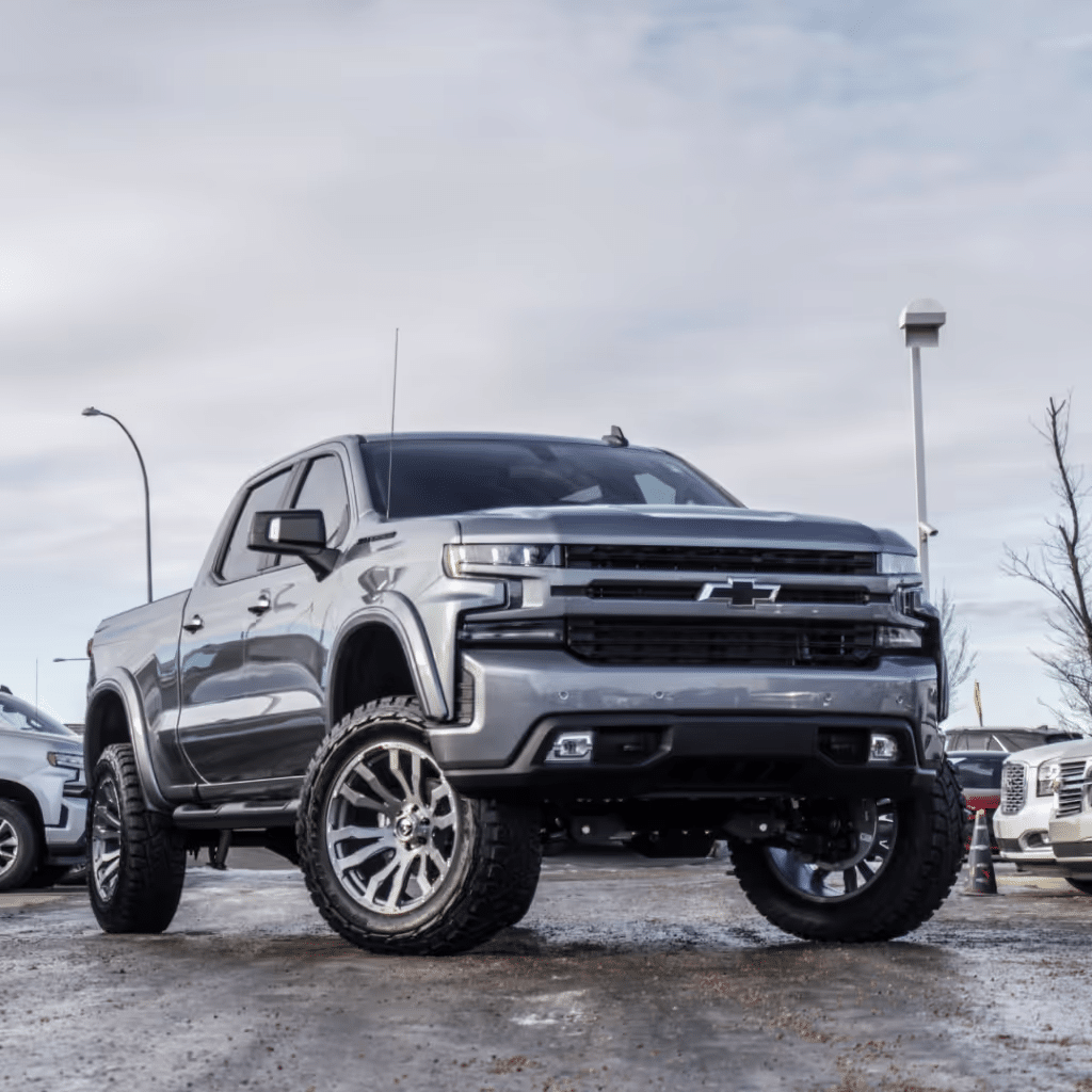Silver Chevrolet truck with oversized wheels and a lifted suspension parked in a dealership lot, showcasing its rugged design against a backdrop of cloudy skies and other vehicles.