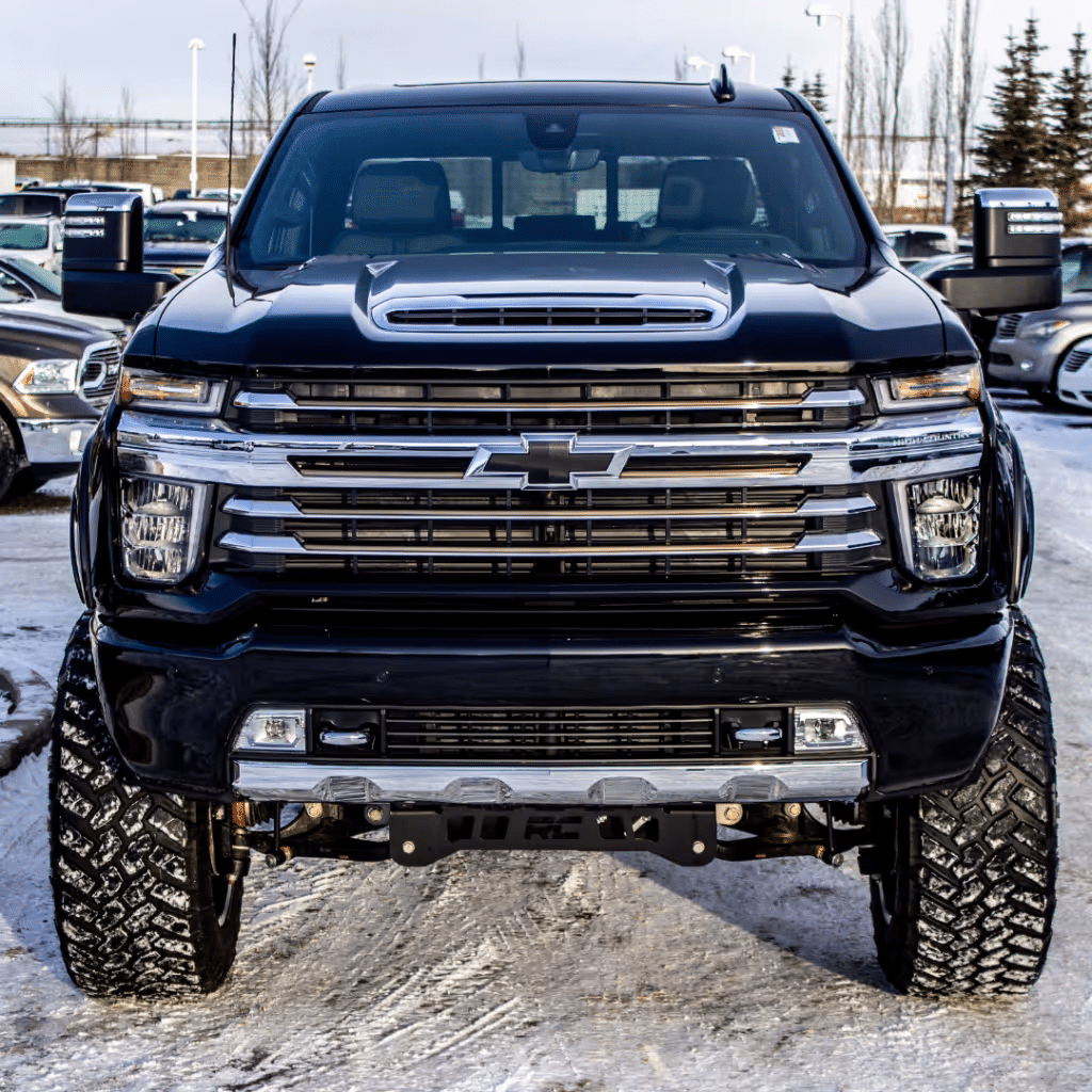 Front view of a black Chevrolet truck with a bold grille and lifted suspension, showcasing aggressive tires and a dynamic presence in a snowy outdoor setting, highlighting its rugged design and off-road capabilities.