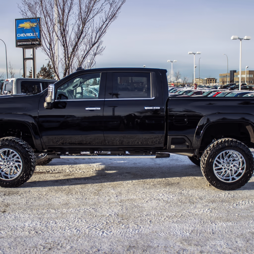 Black pickup truck displayed in a snowy dealership lot with a Chevrolet sign in the background, showcasing stylish chrome wheels and rugged build.