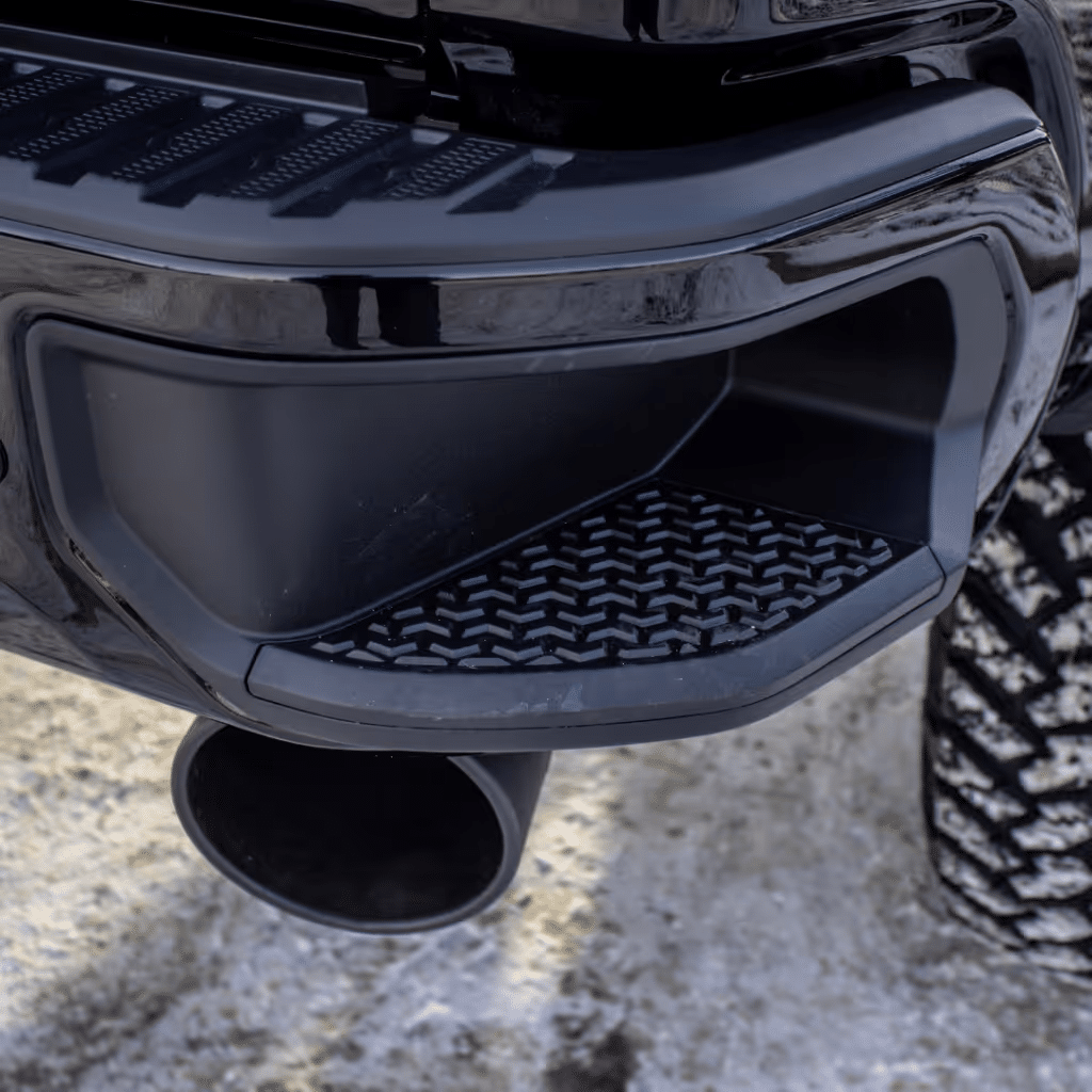 Black truck's rear bumper with a textured step and dual exhaust tip, showcasing a rugged design on a snowy surface.