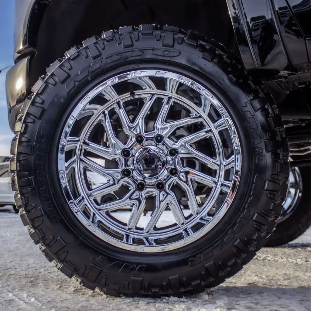 Close-up of a chrome alloy wheel paired with a rugged all-terrain tire, designed for enhanced performance and durability on off-road surfaces. The shiny finish contrasts with the textured tire tread, illustrating a blend of style and functionality.