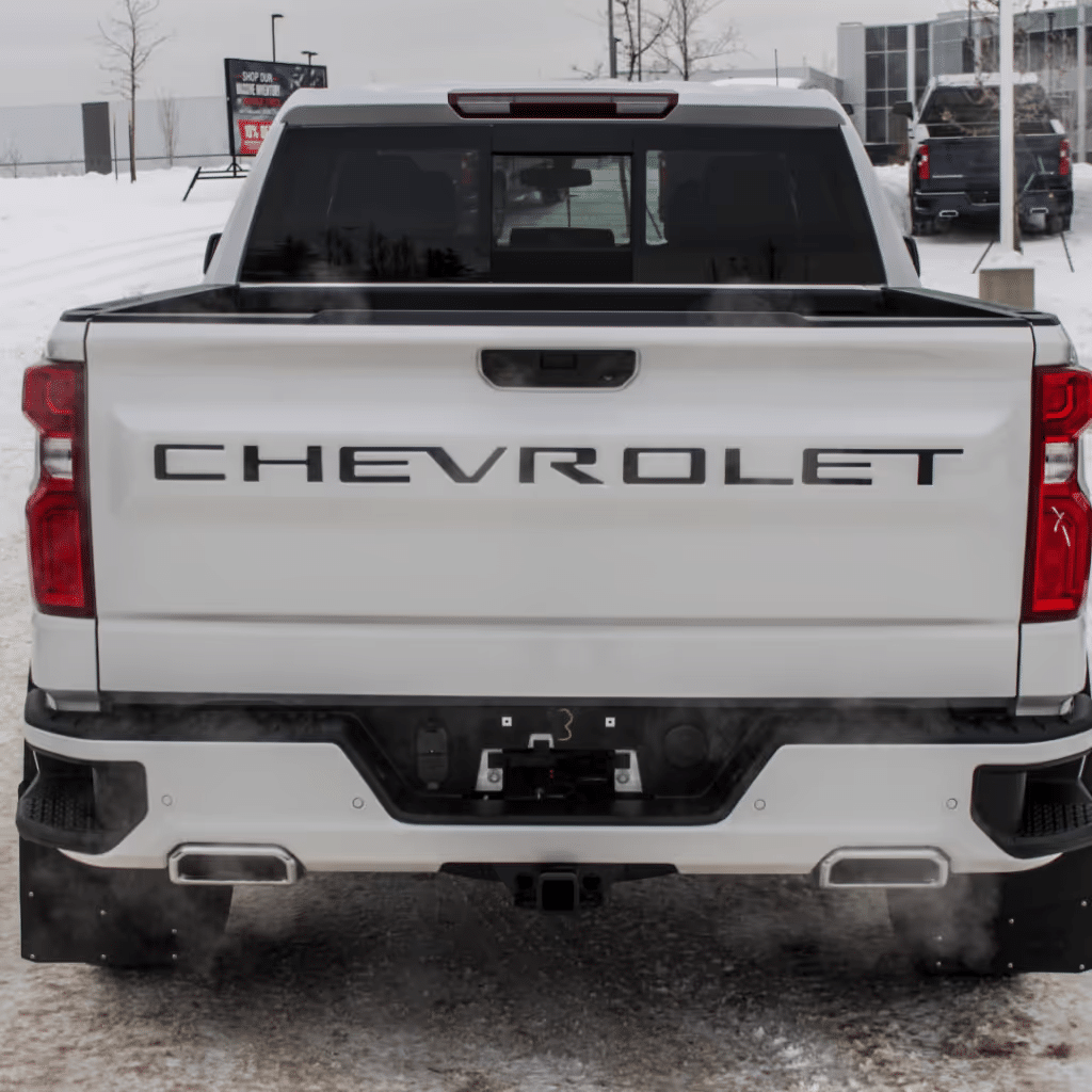 White Chevrolet pickup truck viewed from the rear, showcasing the bold Chevrolet branding on the tailgate, set against a snowy landscape. The truck features a modern design with dual exhaust tips, and faint exhaust smoke is visible, indicating it is running in cold weather conditions.