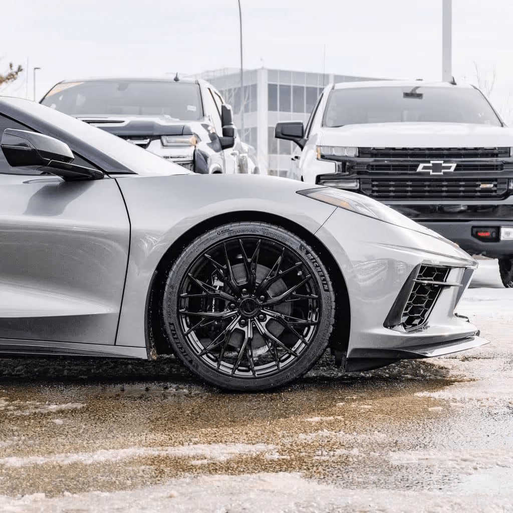 Silver Chevrolet Corvette C8 parked on a wet surface with polished black alloy wheels, showcasing its sleek design and modern features, while other vehicles are visible in the background.