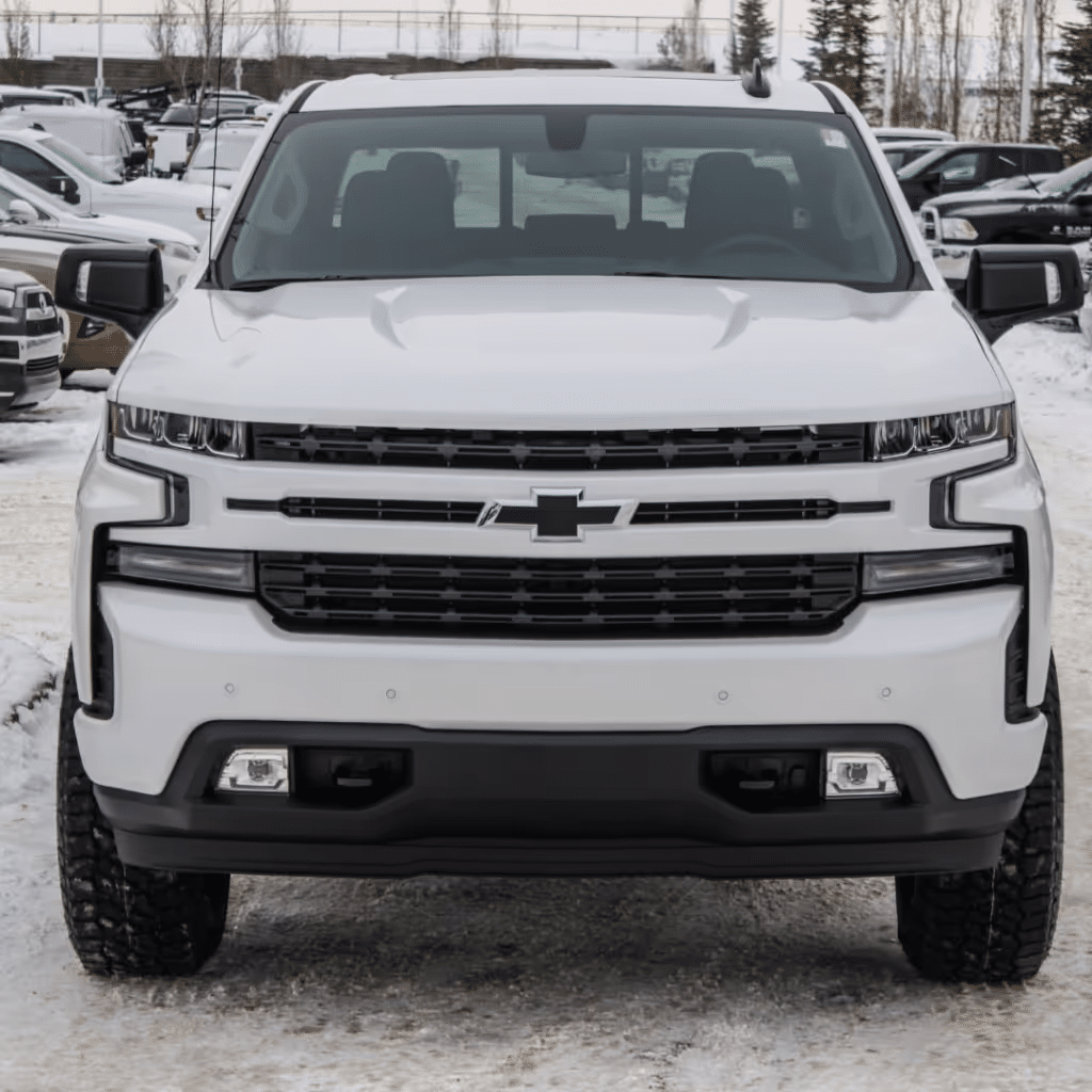 Front view of a white Chevrolet Silverado pickup truck showcasing its bold grille, sleek design, and rugged tires, parked in a snowy outdoor setting with other vehicles in the background.