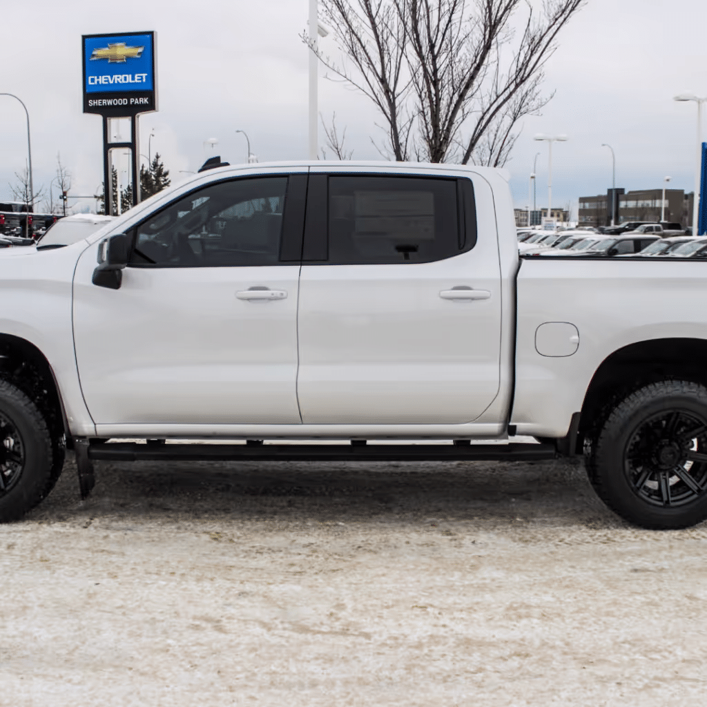 White Chevrolet truck parked at a dealership on a snowy day, showcasing its sleek design and black wheels against a winter backdrop.