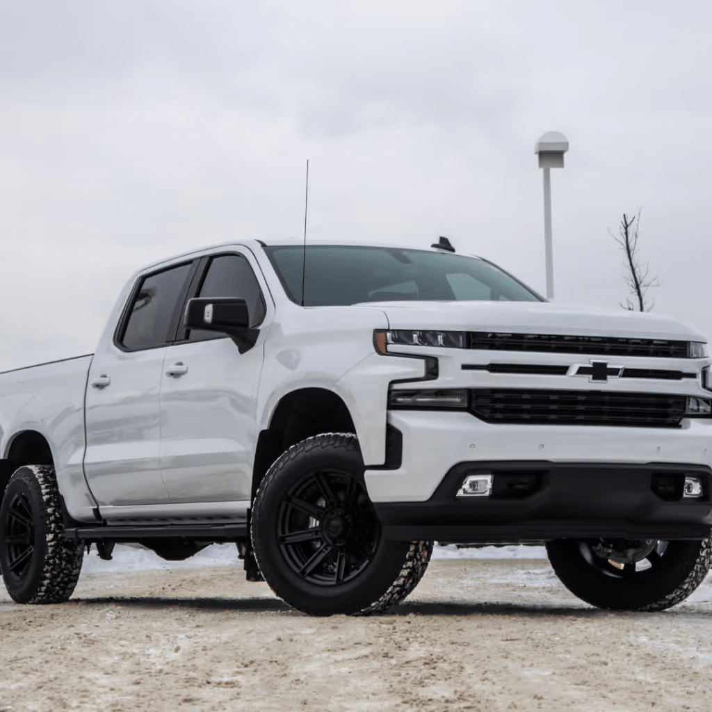 White Chevrolet Silverado pickup truck with black wheels parked on a snowy surface, showcasing its rugged design and modern features against a cloudy sky.