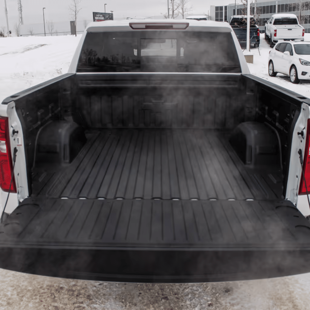 Open truck bed with a black rubber liner, showing steam rising in a snowy environment. The truck is parked in a dealership lot with several vehicles visible in the background.