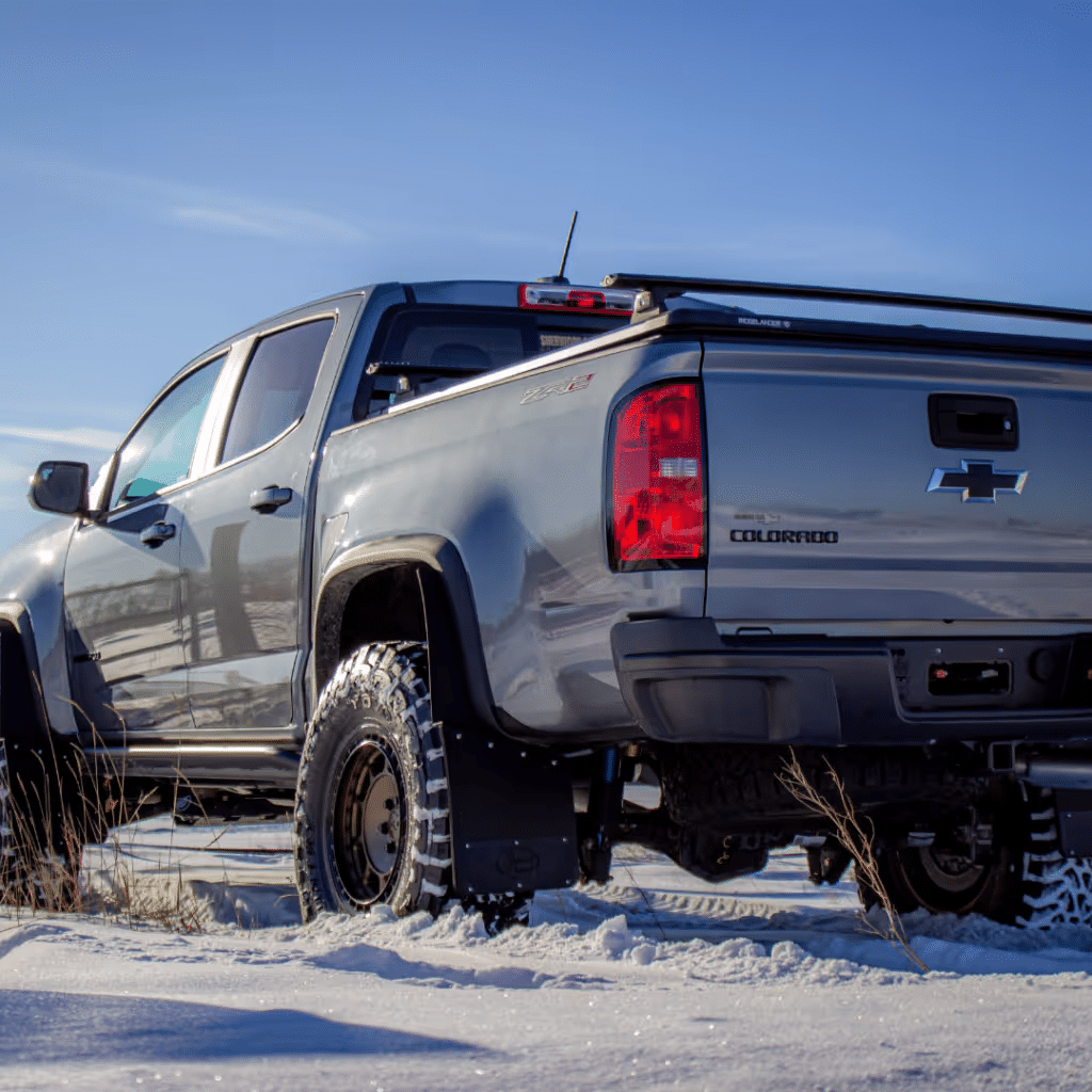 Chevrolet Colorado pickup truck parked in snowy terrain, showcasing rugged tires and a sleek gray exterior under a clear blue sky.
