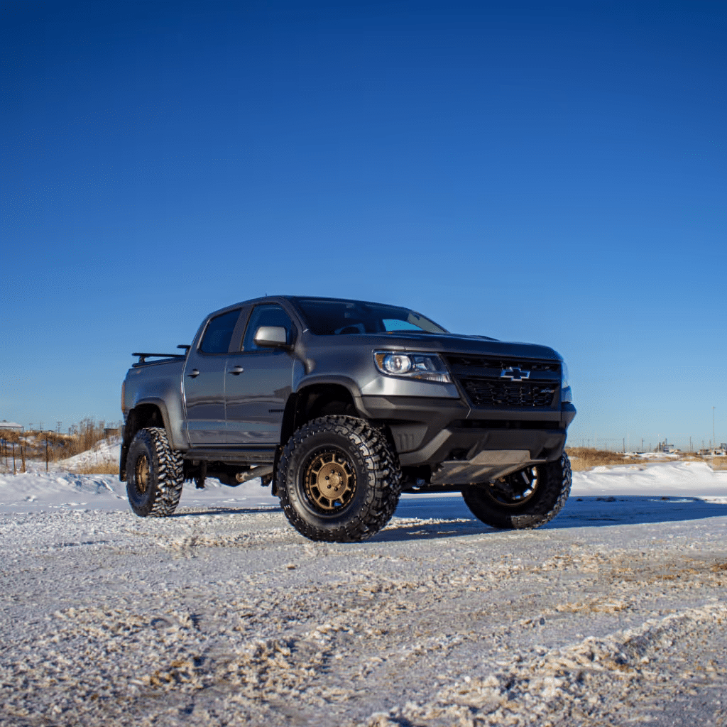 Gray lifted pickup truck parked on snow-covered terrain under a clear blue sky, showcasing rugged tires and an aggressive stance. Ideal for off-road enthusiasts and winter driving.