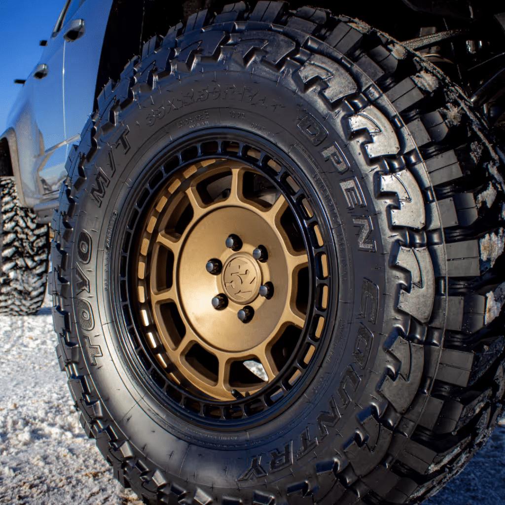 Close-up of a rugged all-terrain tire mounted on a gold alloy wheel, showcasing the tire's aggressive tread design, ideal for off-road performance. The image highlights the tire brand "Toyo" and model "Open Country," emphasizing its durability and traction for adventurous driving.