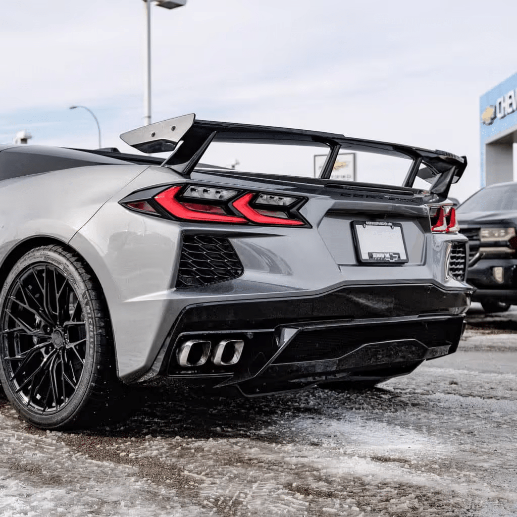 Rear view of a gray Chevrolet Corvette C8 showcasing a prominent rear wing, sleek taillights, and custom black alloy wheels, parked outdoors likely at a dealership with snowy ground and a clear sky.