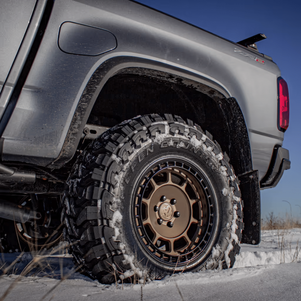 Close-up of a rugged off-road tire on a gray pickup truck, showcasing detailed tread patterns and a stylish bronze wheel, set against a snowy landscape. Ideal for winter driving and off-road adventures.