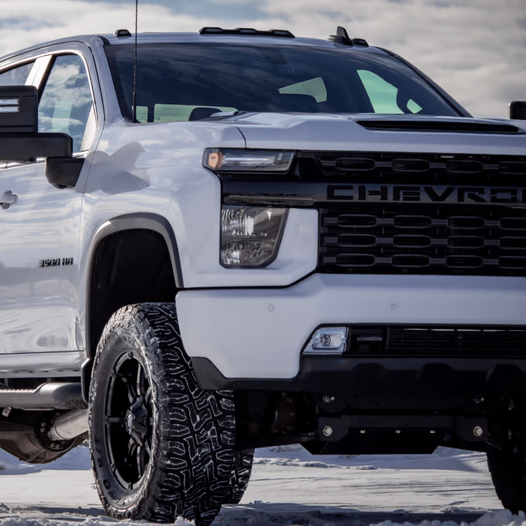 White Chevrolet Silverado 3500 HD truck parked on snow, showcasing its robust build, black accents, and off-road tires. The image highlights the truck's front view with modern features and a sleek design against a cloudy sky backdrop.