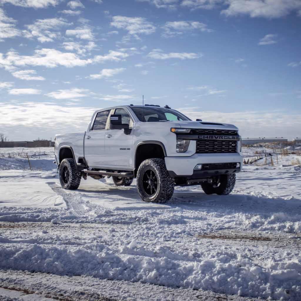 White Chevrolet Silverado 2500HD pickup truck with off-road tires parked on snowy terrain under a blue sky with scattered clouds.