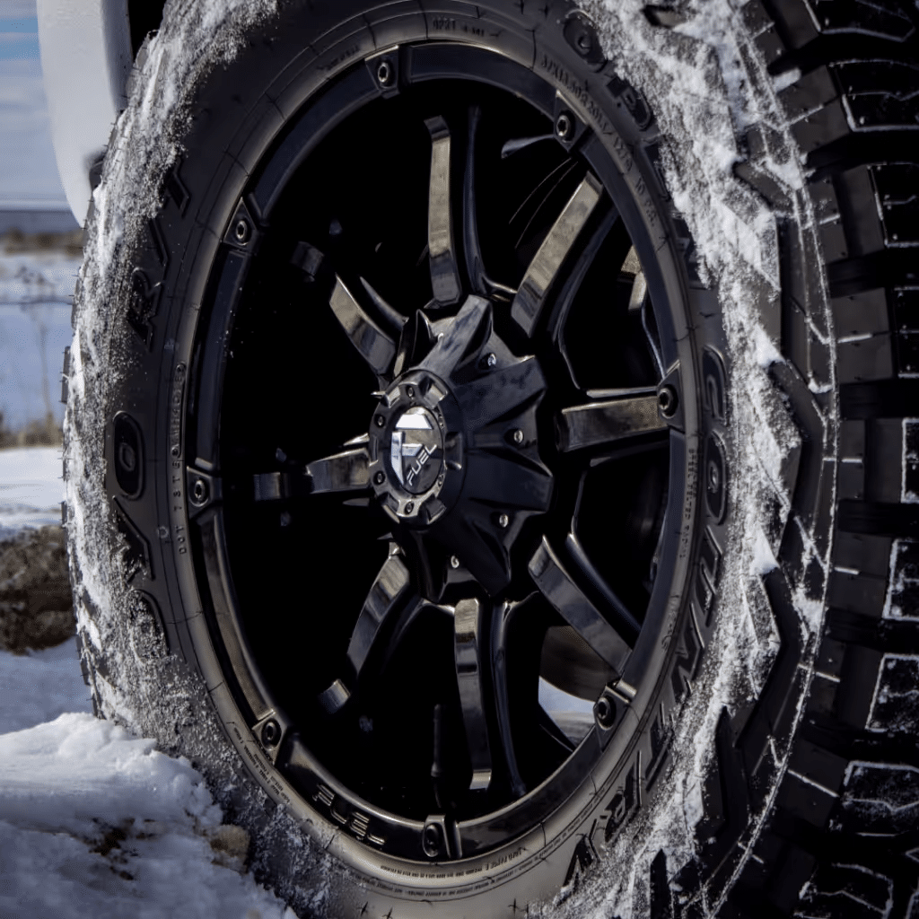 Close-up of a stylish black alloy wheel with rugged all-terrain tire, partially covered in snow, showcasing its design and traction capabilities in winter conditions.