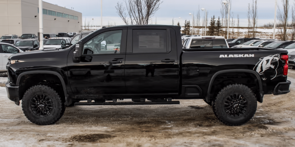 Black Alaskan edition pickup truck with oversized tires and black alloy wheels, parked in a snowy lot surrounded by other vehicles. The truck features a stylish design with a prominent logo on the side and is ideal for off-road adventures.