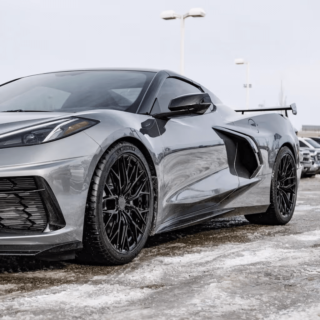 Image of a sleek silver Corvette C8 parked on a snowy surface, showcasing its modern aerodynamic design and black alloy wheels, with a dealership in the background.