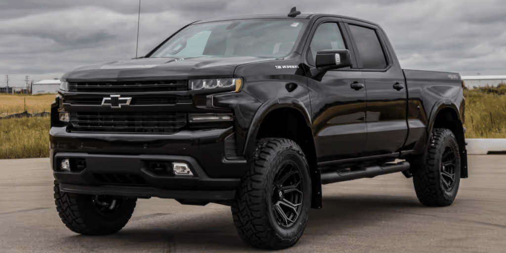 Black Chevrolet Silverado 1500 with off-road tires and a lifted suspension, parked on an outdoor lot under cloudy skies. The truck features a modern design with angular headlights and bold front grille, showcasing its rugged appeal and capability.