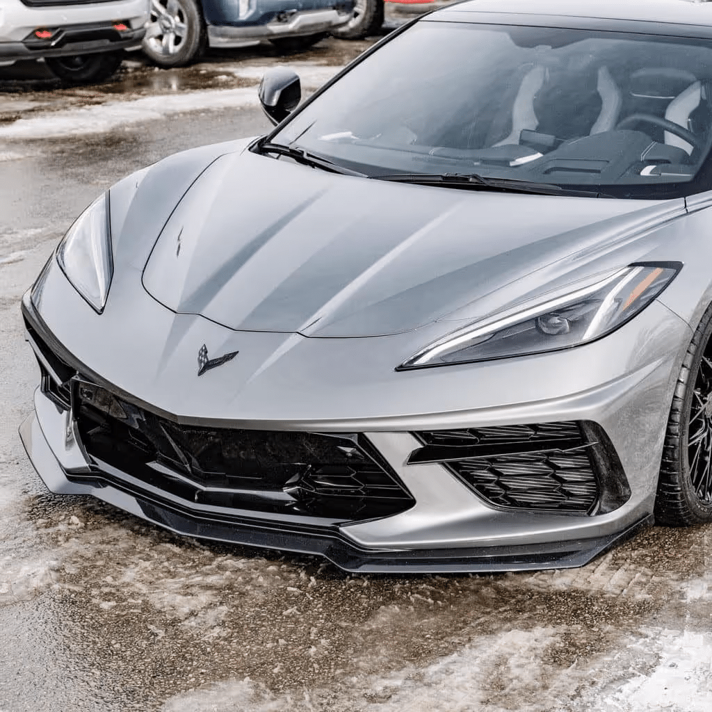 Silver Chevrolet Corvette C8 parked on a snowy surface, showcasing its sleek aerodynamic design and aggressive front grille. The image captures the headlights and distinctive sculpted body lines, highlighting the car's performance-oriented features.
