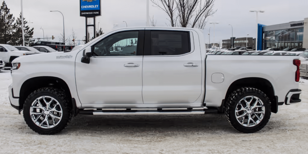 Side view of a white Chevrolet Silverado pickup truck parked in a snowy dealership lot, showcasing its polished alloy wheels and rugged design, with a Chevrolet dealership in the background.