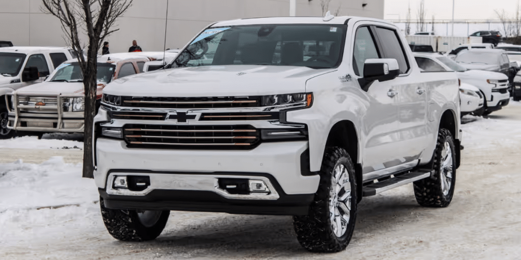 White Chevrolet Silverado pickup truck parked in a snowy dealership lot, showcasing its modern design with chrome accents and off-road tires.